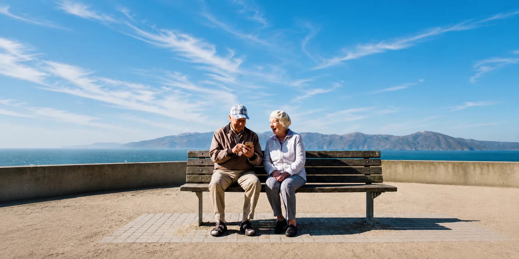 An older couple smiling at a phone together on a bench by the water